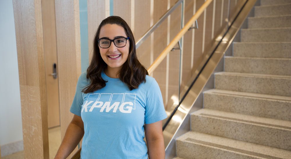 A female student wearing a blue KPMG t-shirt smiles standing near the steps of the Business building lobby